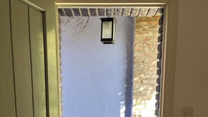 Une porte-fenêtre donnant sur une terrasse en bois, d’où l’on aperçoit un mur de briques et une petite fenêtre.