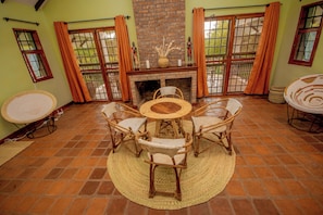 A room with a brick fireplace, a round table with chairs, and a view through large windows with orange curtains. - Amani Farmhouse (Arusha)