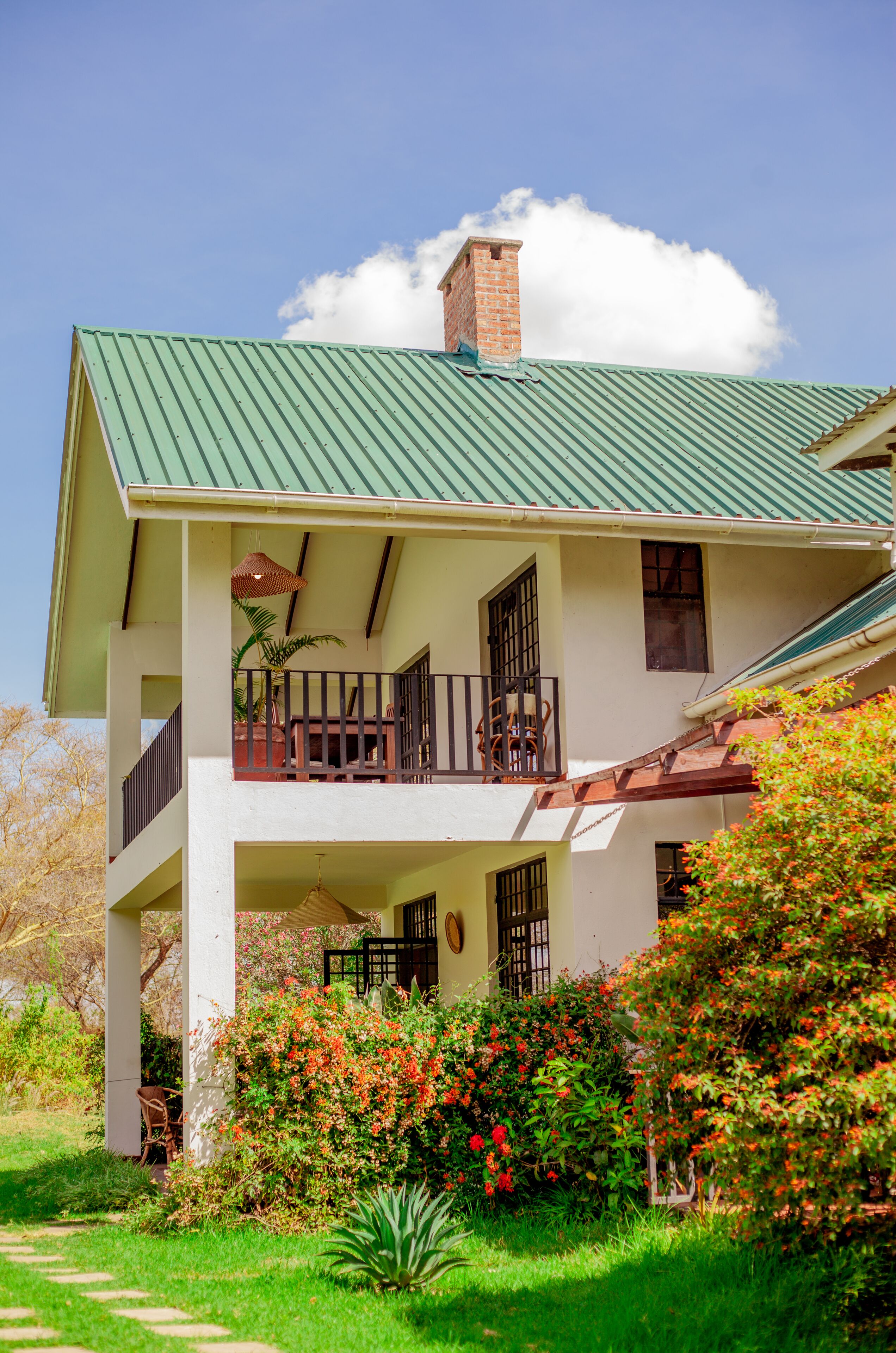 A two-story building with a green metal roof, a balcony with a railing, and a garden with red flowers.
