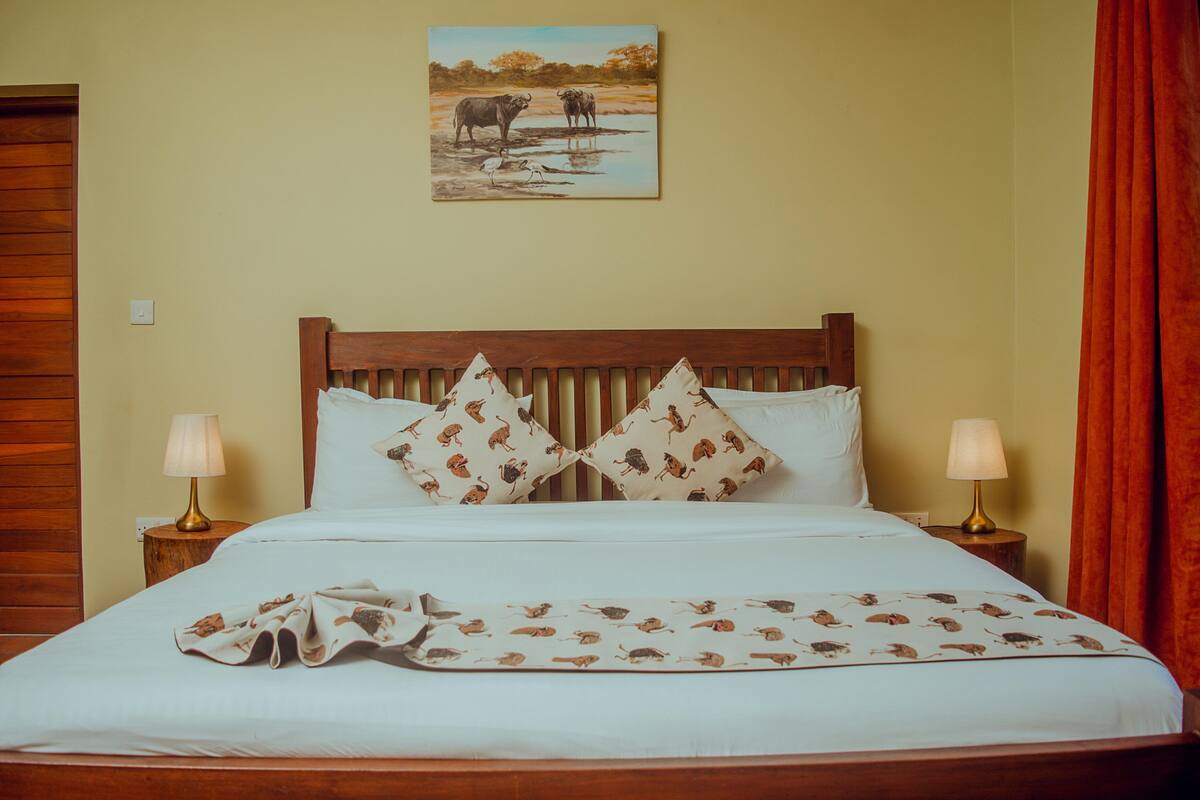 Ein Bett mit einem gemusterten Überwurf, zwei Kissen mit Tiermotiven, ein Wandbild mit Wildtieren und zwei Nachttischlampen.