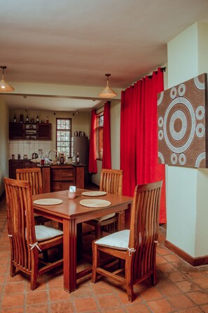A dining area with a wooden table and chairs, a kitchen in the background, and a red curtain. - Amani Farmhouse (Arusha)