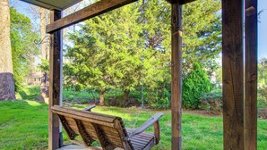 Un banc-balançoire en bois, placé sous une pergola en bois, offre une vue sur une zone herbeuse et des arbres.