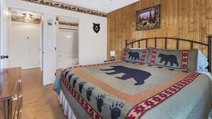 Une chambre avec un mur en bois, un lit recouvert d’une couette à motifs d’ours, un ventilateur de plafond et une porte donnant sur une autre pièce.