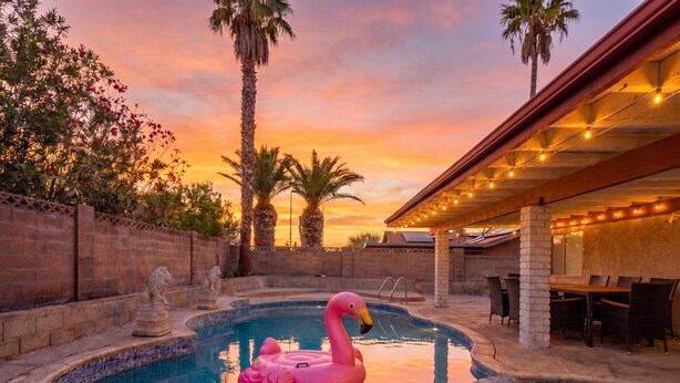 En pool med en rosa uppblåsbar flamingofigur, palmer och en täckt uteplats.