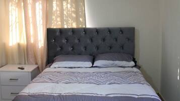 A bedroom with a tufted headboard, a bed with patterned bedding, a nightstand, a ceiling fan, and an air conditioning unit.