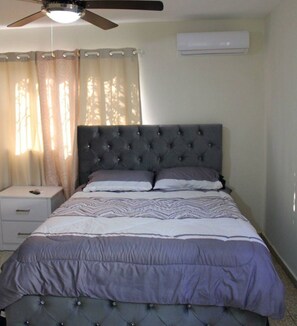 A bedroom with a tufted headboard, a bed with patterned bedding, a nightstand, a ceiling fan, and an air conditioning unit. - Family house near the beach (La Romana)