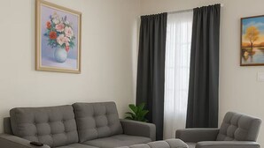 Un salon avec un canapé d’angle gris, deux fauteuils, un ventilateur de plafond et un tableau représentant des fleurs.