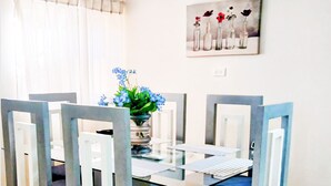 Un coin repas avec une table à plateau en verre, entourée de chaises à la structure grise et aux coussins foncés. Un vase avec des fleurs bleues est posé sur la table.