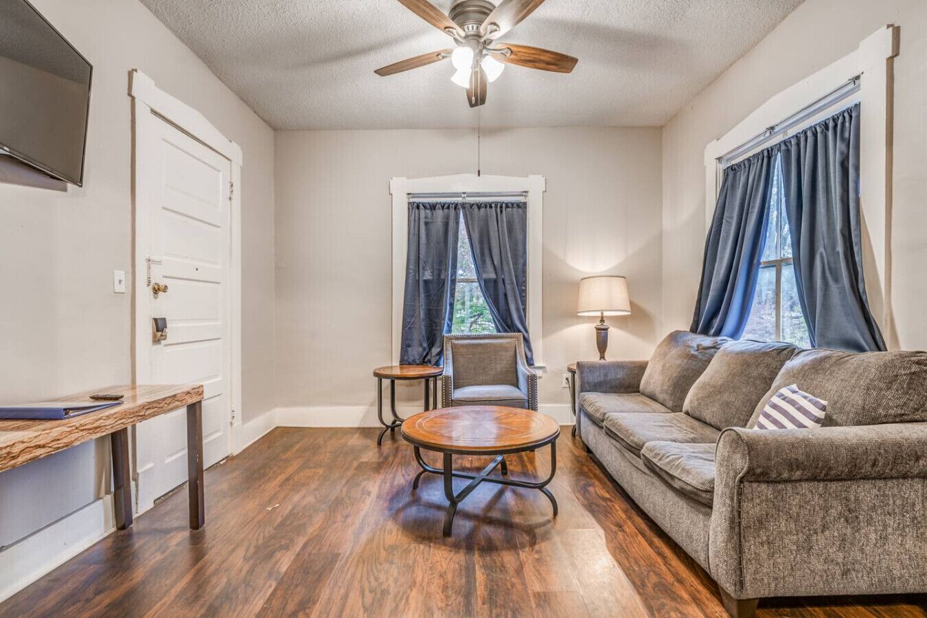 A living room with a wooden floor, a ceiling fan, a sofa, a coffee table, and a TV mounted on the wall.