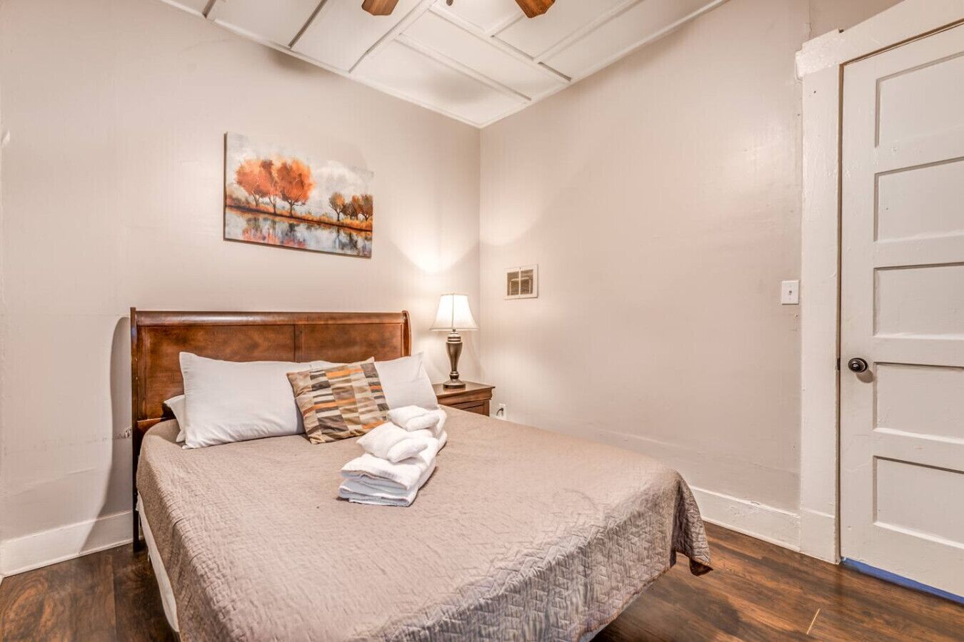 A neatly made bed with a wooden headboard, a painting above it, and a nightstand with a lamp.