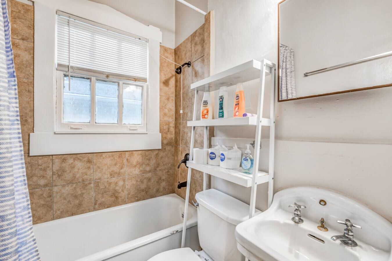 Ein Badezimmer mit Badewanne, Toilette und Waschbecken. Ein Fenster mit Jalousien sowie Regale mit Pflegeprodukten.