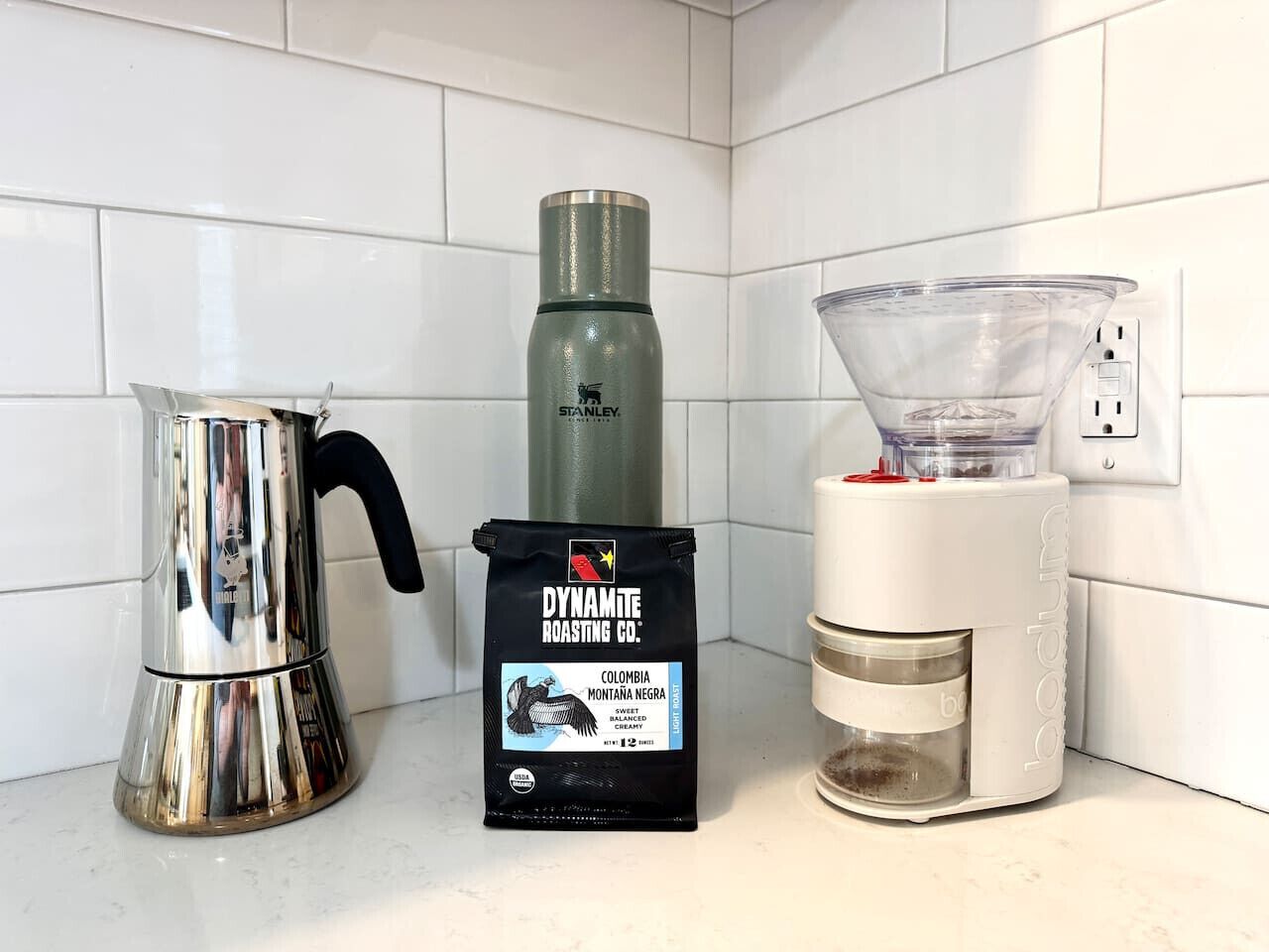 Una encimera de cocina con una cafetera de acero inoxidable, un termo verde, un molinillo de café y un paquete de café Colombia Montaña Negra de Dynamite Roasting Co.