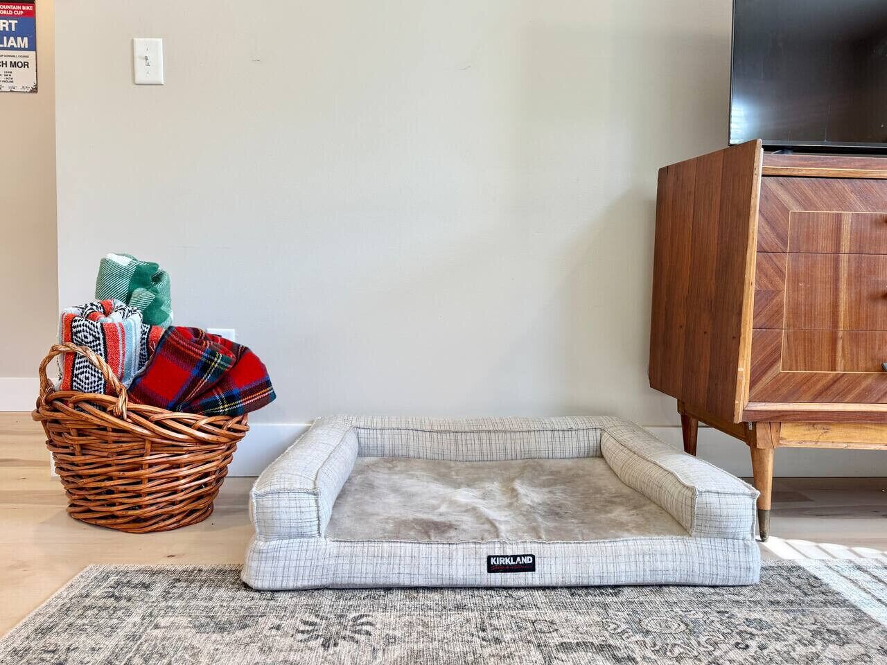 A pet bed with a label, a wicker basket with blankets, and a wooden cabinet.