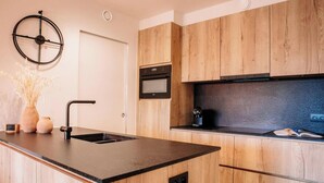 A modern kitchen with wooden cabinets, a black countertop, a built-in oven, and a wall-mounted clock. - SAND–Modern apartment with garage near the beach (Ostend)