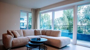 A modern living room with a beige sectional sofa, a round coffee table, and large windows offering a view of greenery and a building. - SAND–Modern apartment with garage near the beach (Ostend)