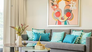 Una sala de estar moderna con un sofá gris, cojines estampados en verde azulado y blanco, una mesa de centro de vidrio y un cuadro abstracto enmarcado en la pared.
