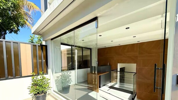 Un espacio exterior moderno con un ventanal de vidrio que da a una cocina, una terraza de madera y una planta en maceta.
