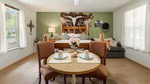 A dining area with a round wooden table, two chairs, a ceiling fan, a bed with a wooden headboard, and a green wall with a decorative piece.