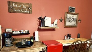 Una cocina con cafetera, pileta y un refrigerador rojo. Hay sillas de madera y una mesa. Un cartel en la pared dice 'Kick off Yer boots Cowboy'.