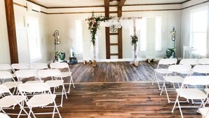 Una sala con sillas plegables blancas dispuestas en filas, suelo de madera y un arreglo floral decorativo al frente.