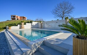 A swimming pool with steps leading into it, surrounded by a landscaped area with a palm tree and a building in the background. - Beautiful home in Banfi (Banfi)