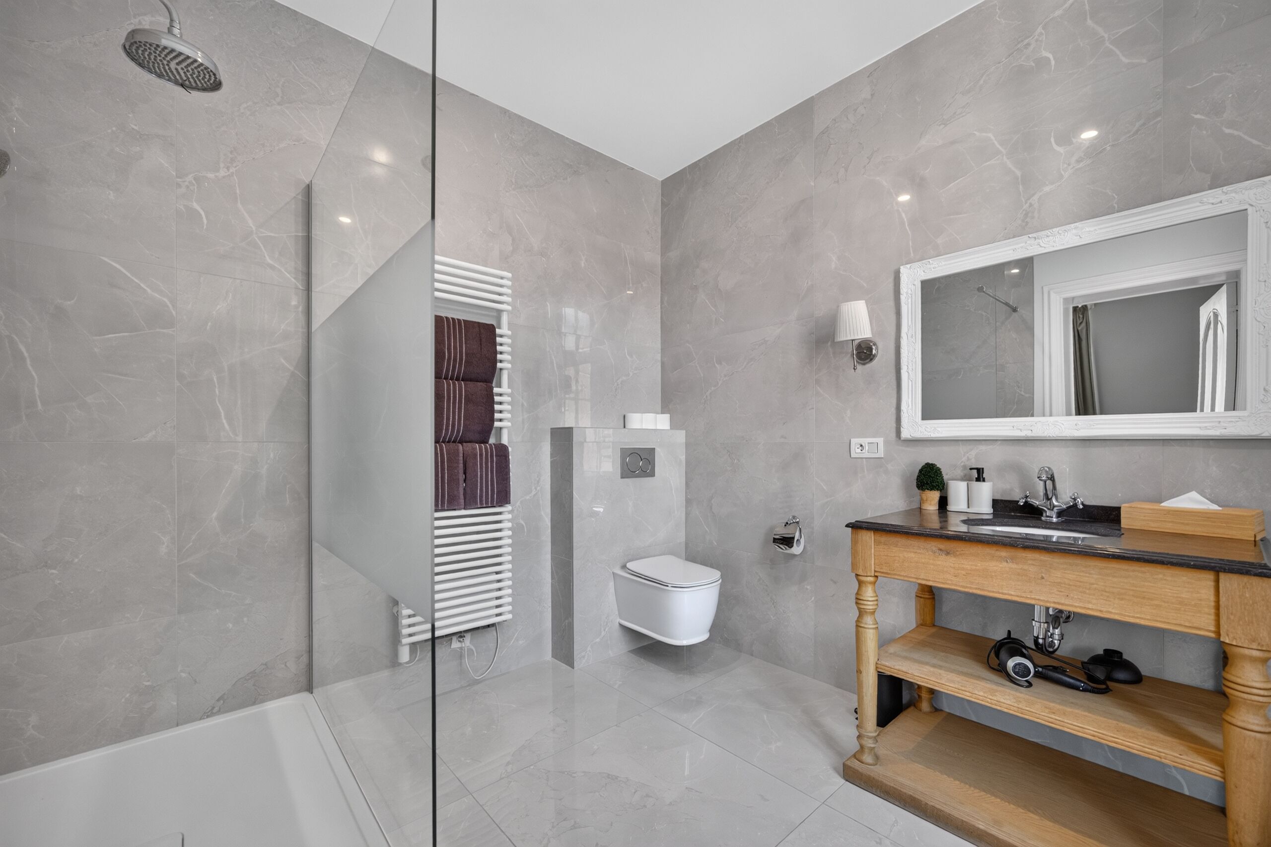 Un baño moderno con cabina de ducha de vidrio, inodoro blanco, tocador de madera con espejo y suelo de baldosas de mármol.