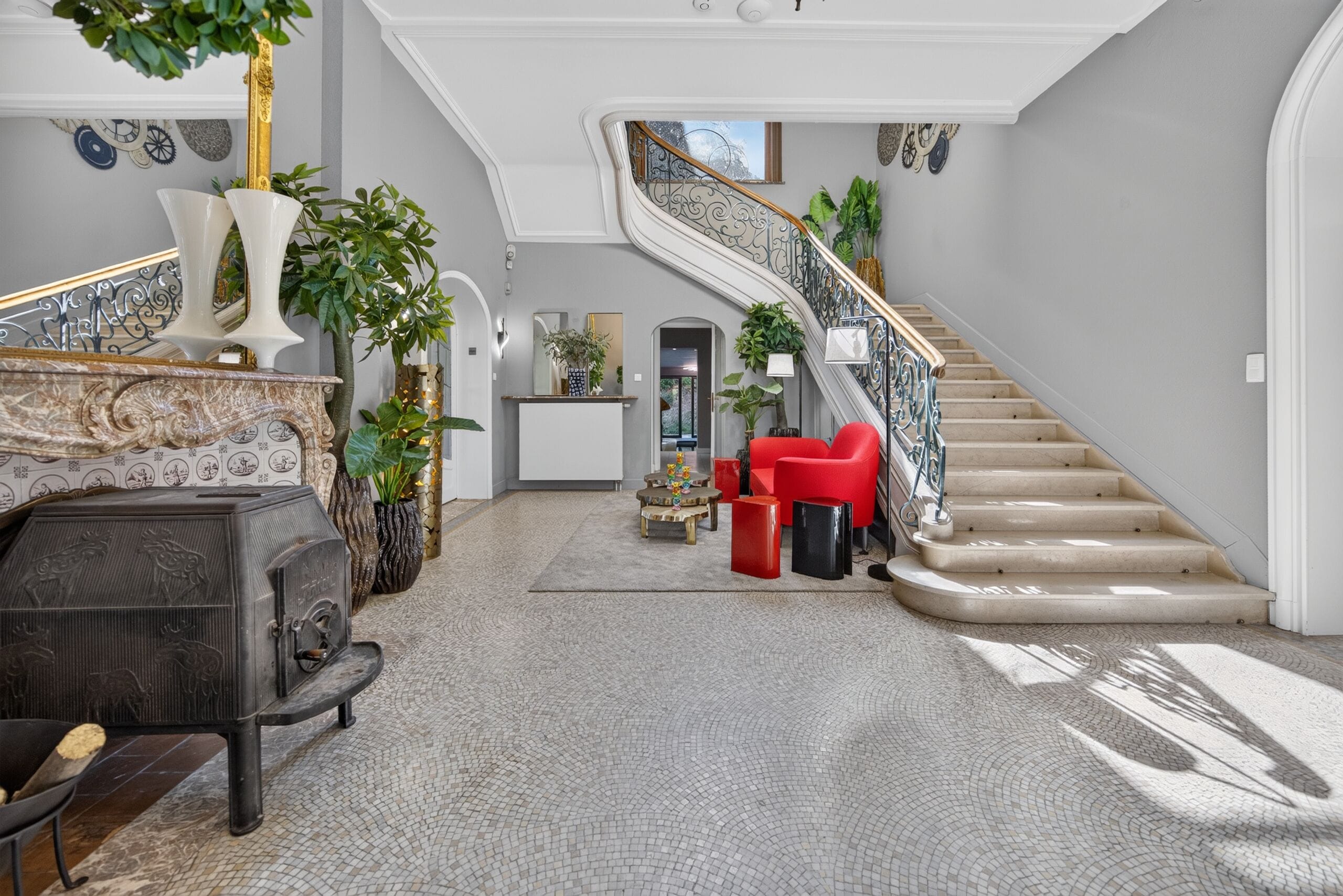 A spacious foyer with a grand staircase, a seating area with red chairs, and a variety of decorative plants.