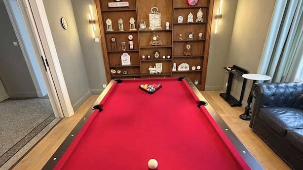 A pool table with a red felt surface, surrounded by a wooden cabinet with clocks and trophies. - Villa Damseaux (Spa)