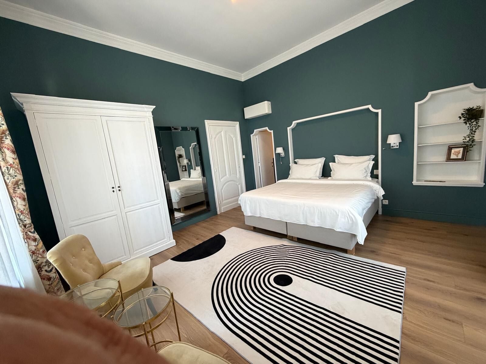 A bedroom with a bed, wardrobe, mirror, bedside table, lamp, and a patterned rug.