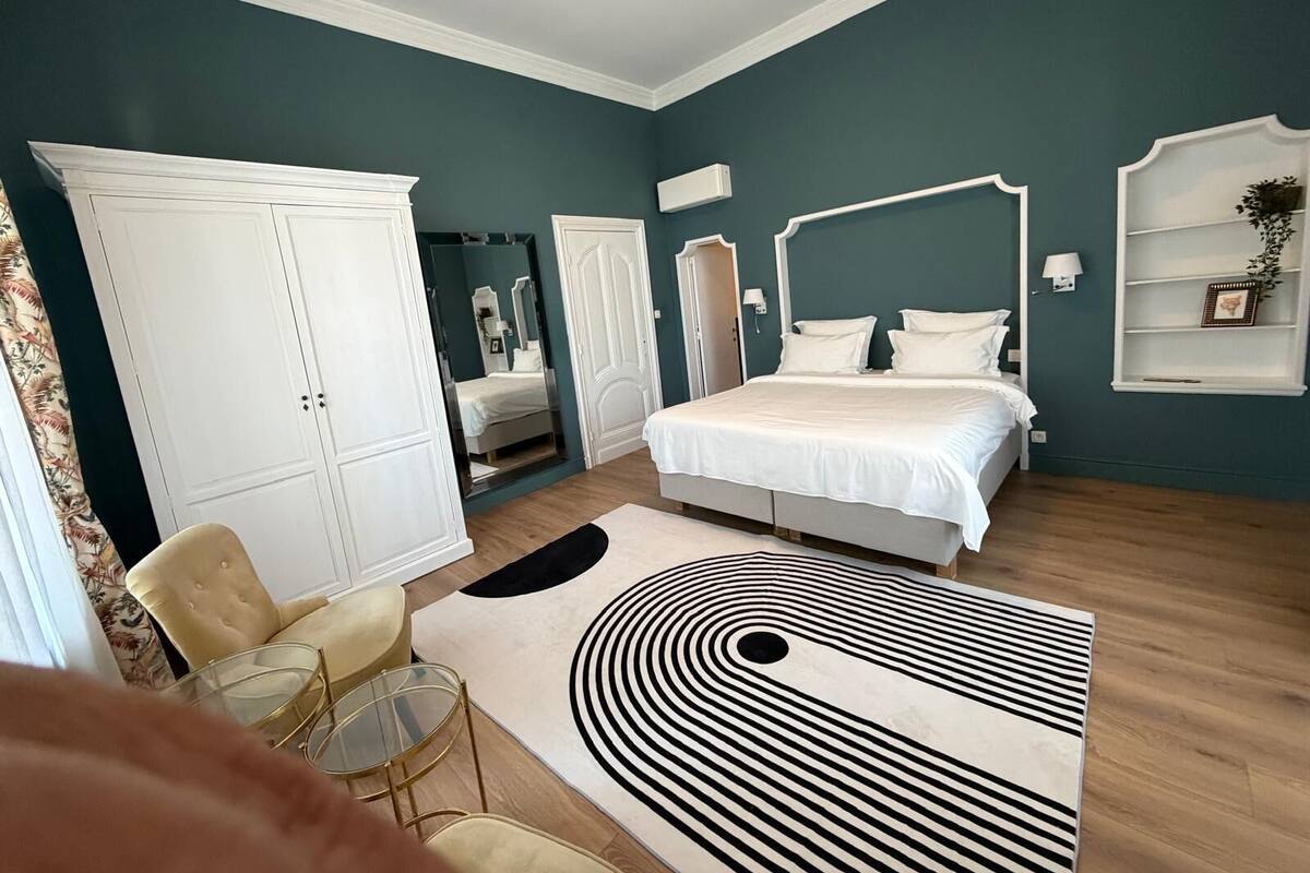 A bedroom with a bed, wardrobe, mirror, bedside table, lamp, and a patterned rug.