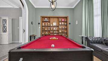 A pool table with a red felt surface, surrounded by a wooden cabinet with clocks, a leather sofa, and a billiards cue rack.