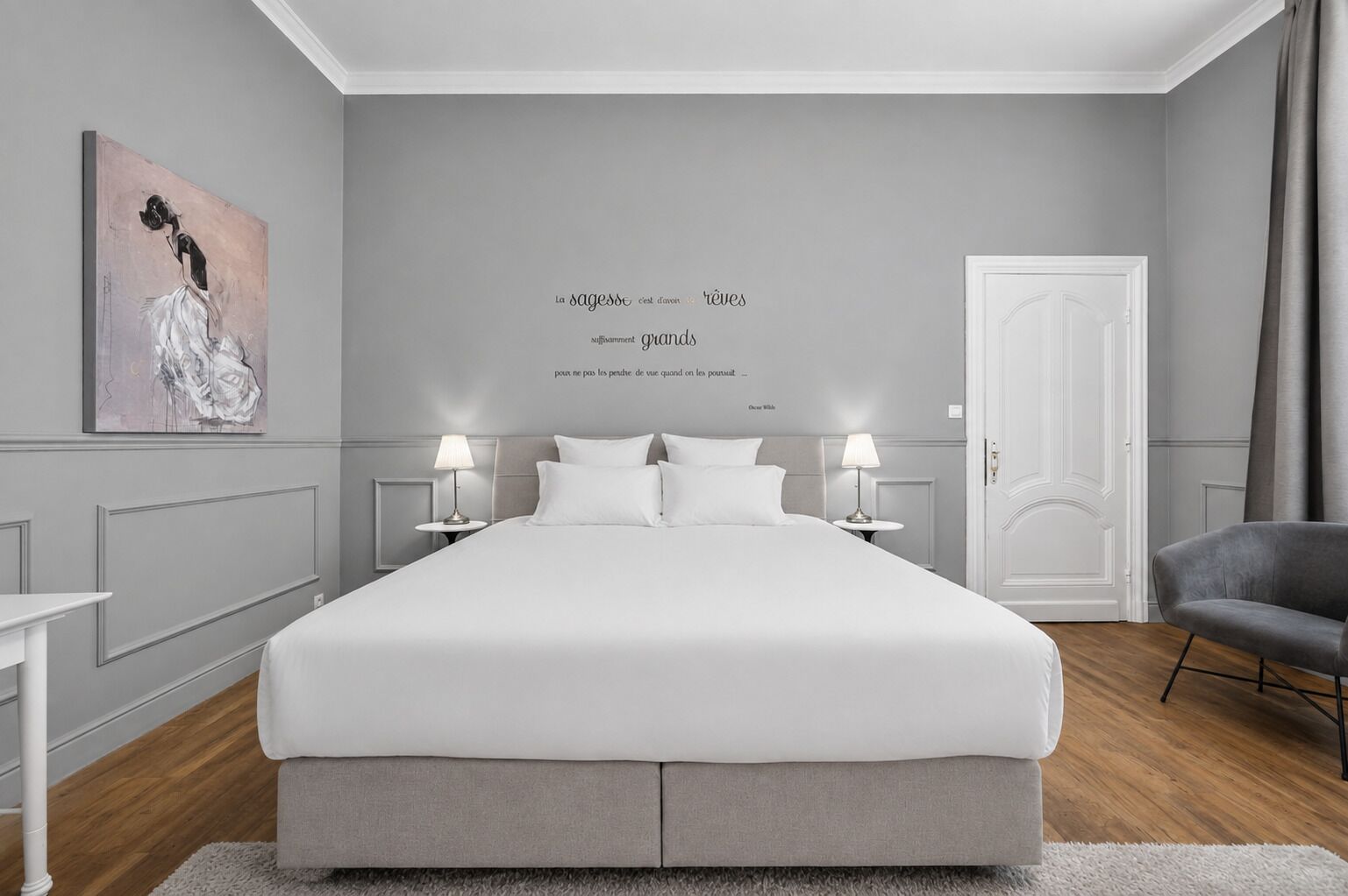 Un dormitorio moderno con una cama grande, dos mesitas de noche con lámparas, una silla gris y una puerta.