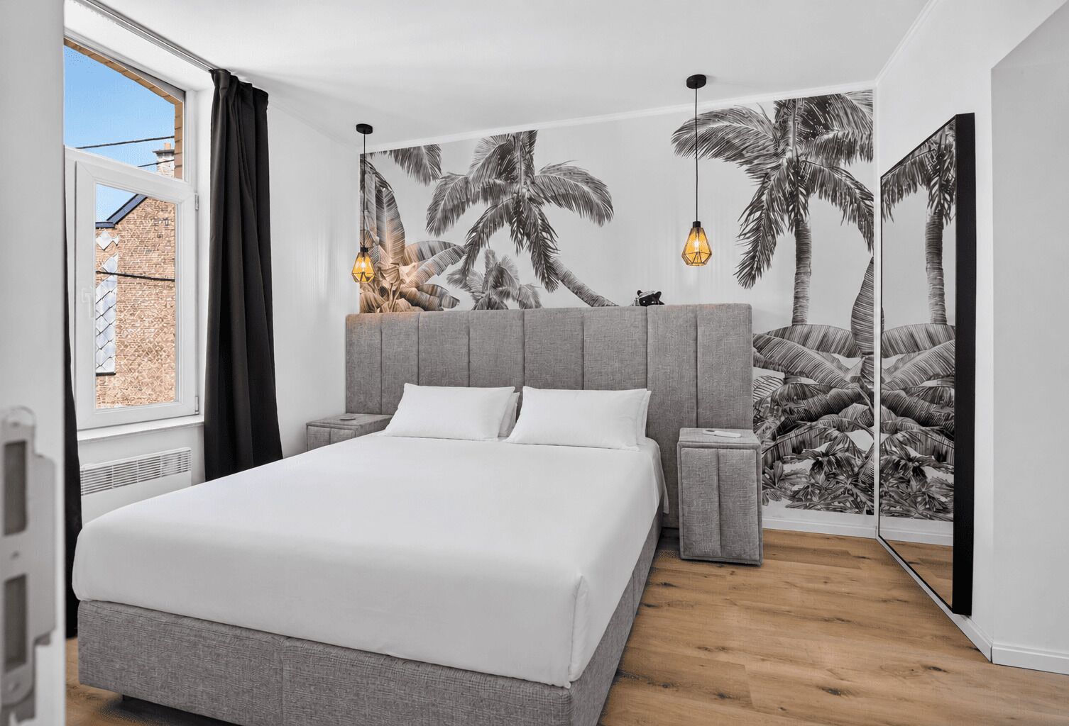 A bedroom with a large bed, a grey headboard, bedside tables, and a wall with a tropical leaf pattern.