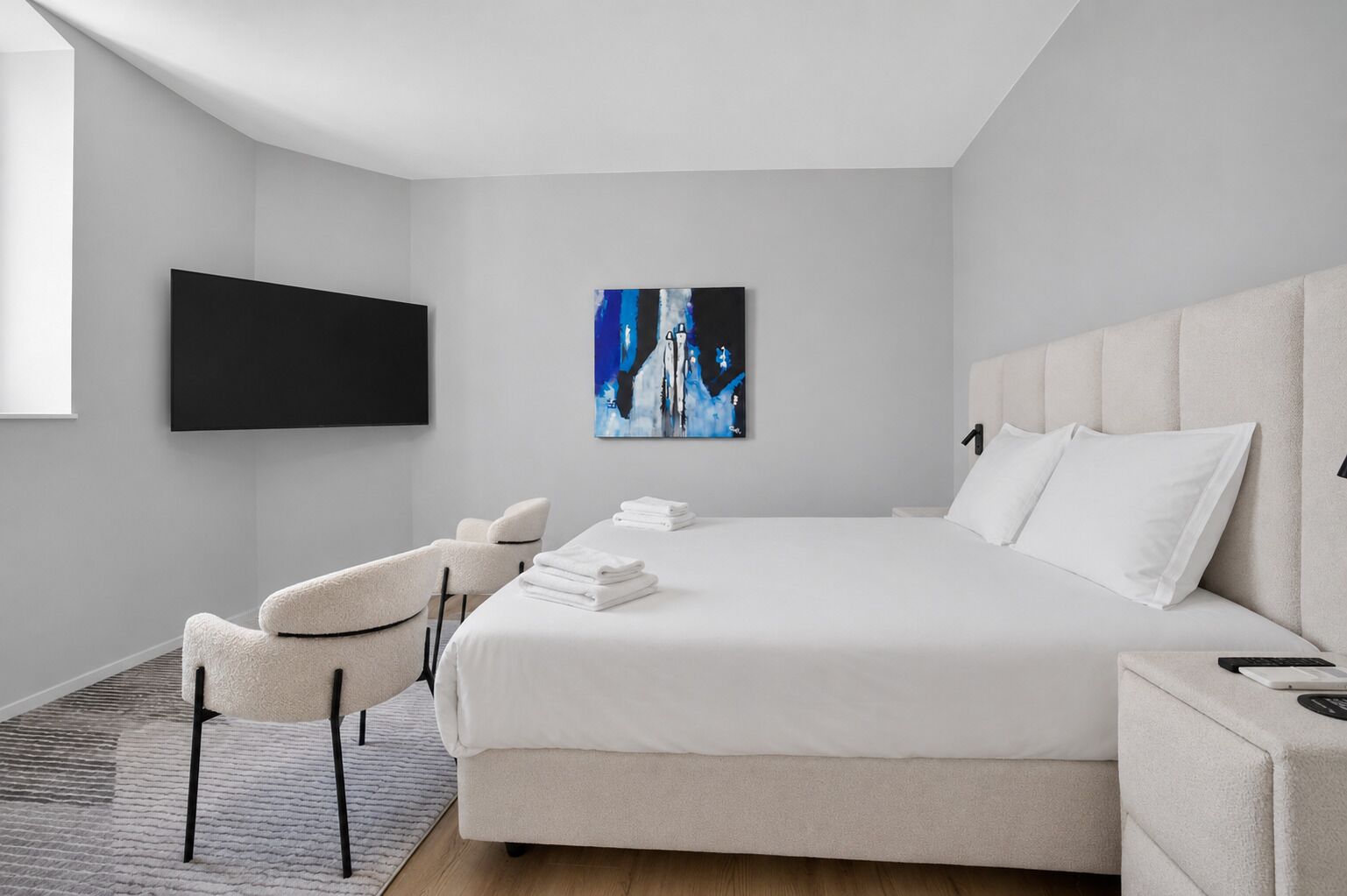 Une chambre d’hôtel moderne dotée d’un grand lit, d’un téléviseur à écran plat, d’un tableau et d’un coin salon avec un fauteuil et une petite table.