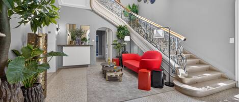 A spacious living area with a red sofa, a staircase with ornate railings, and a large potted plant.