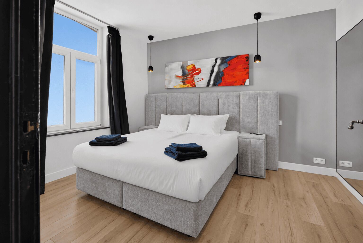 A modern bedroom with a large bed, a grey headboard, a bedside table, a wall art piece, and a window with curtains.