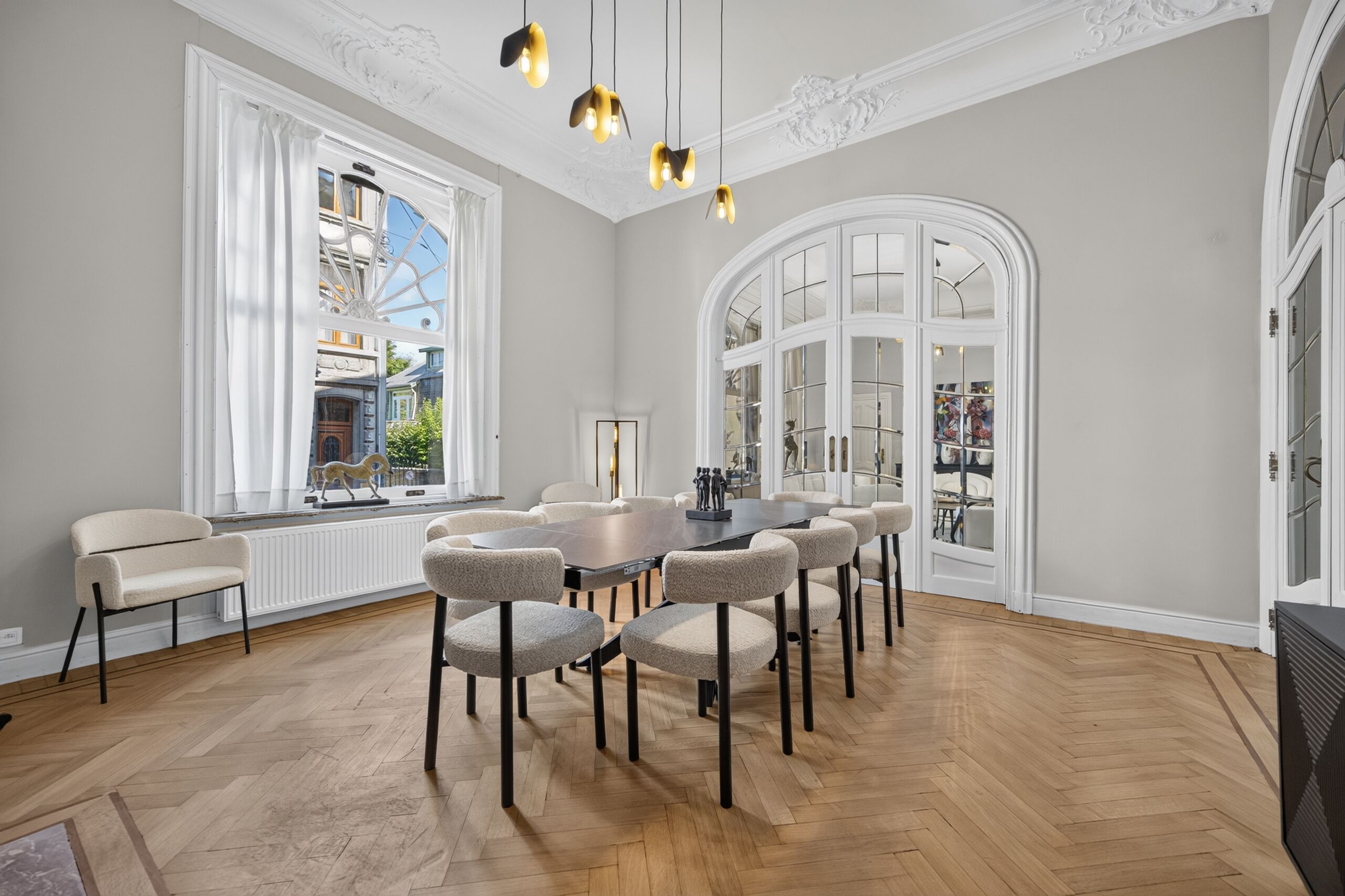 A spacious room with a large dining table surrounded by chairs, large windows with white curtains, and a decorative ceiling.