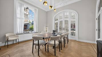 A spacious room with a large dining table surrounded by chairs, large windows with white curtains, and a decorative ceiling.