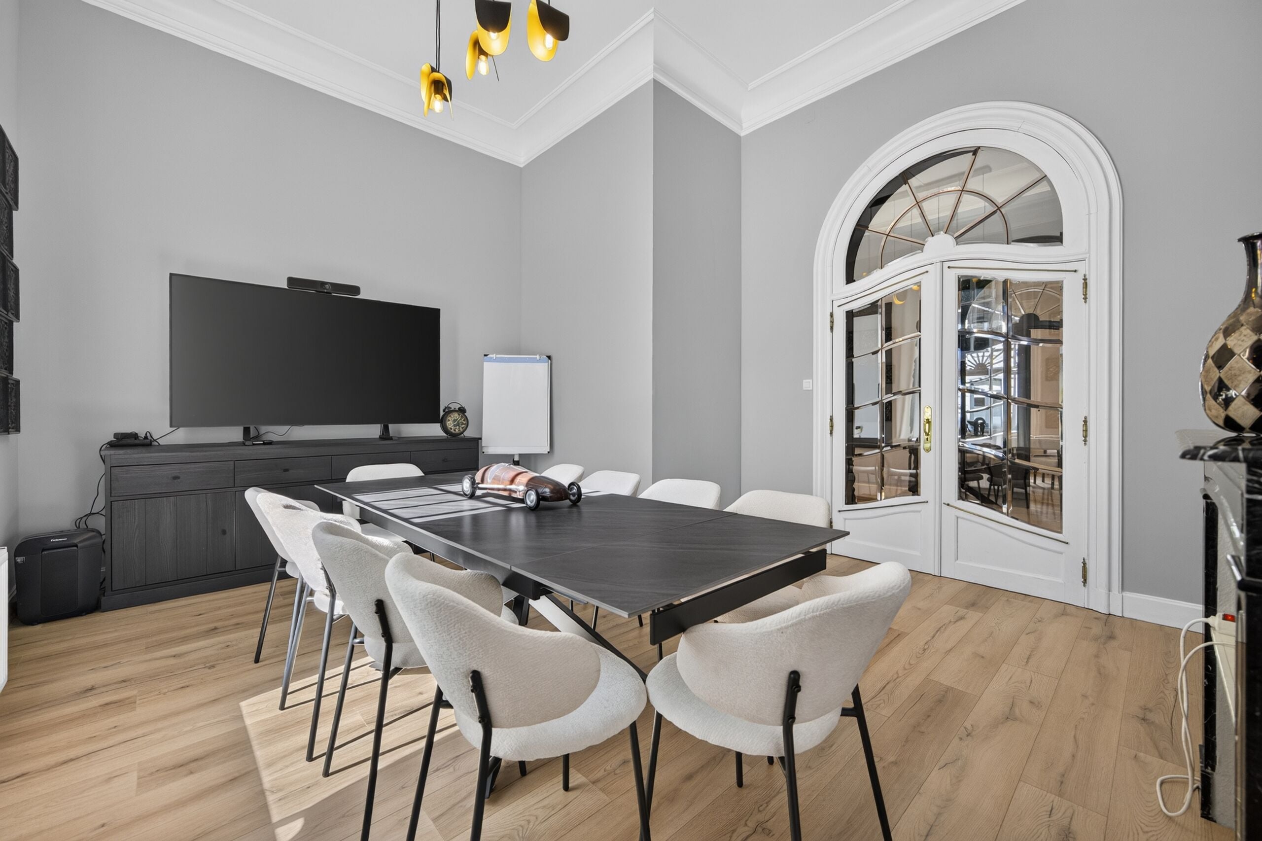 Une salle de réunion moderne, dotée d’une grande table, de chaises blanches, d’un téléviseur à écran plat et d’un vase décoratif.