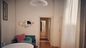 Een kamer met een ronde eettafel, een bank met kussens, een spiegel aan de muur en een gang naar een andere kamer.