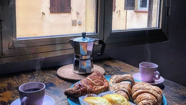 Een tafel met gebak en koffiekopjes bij een raam met uitzicht op een gebouw.