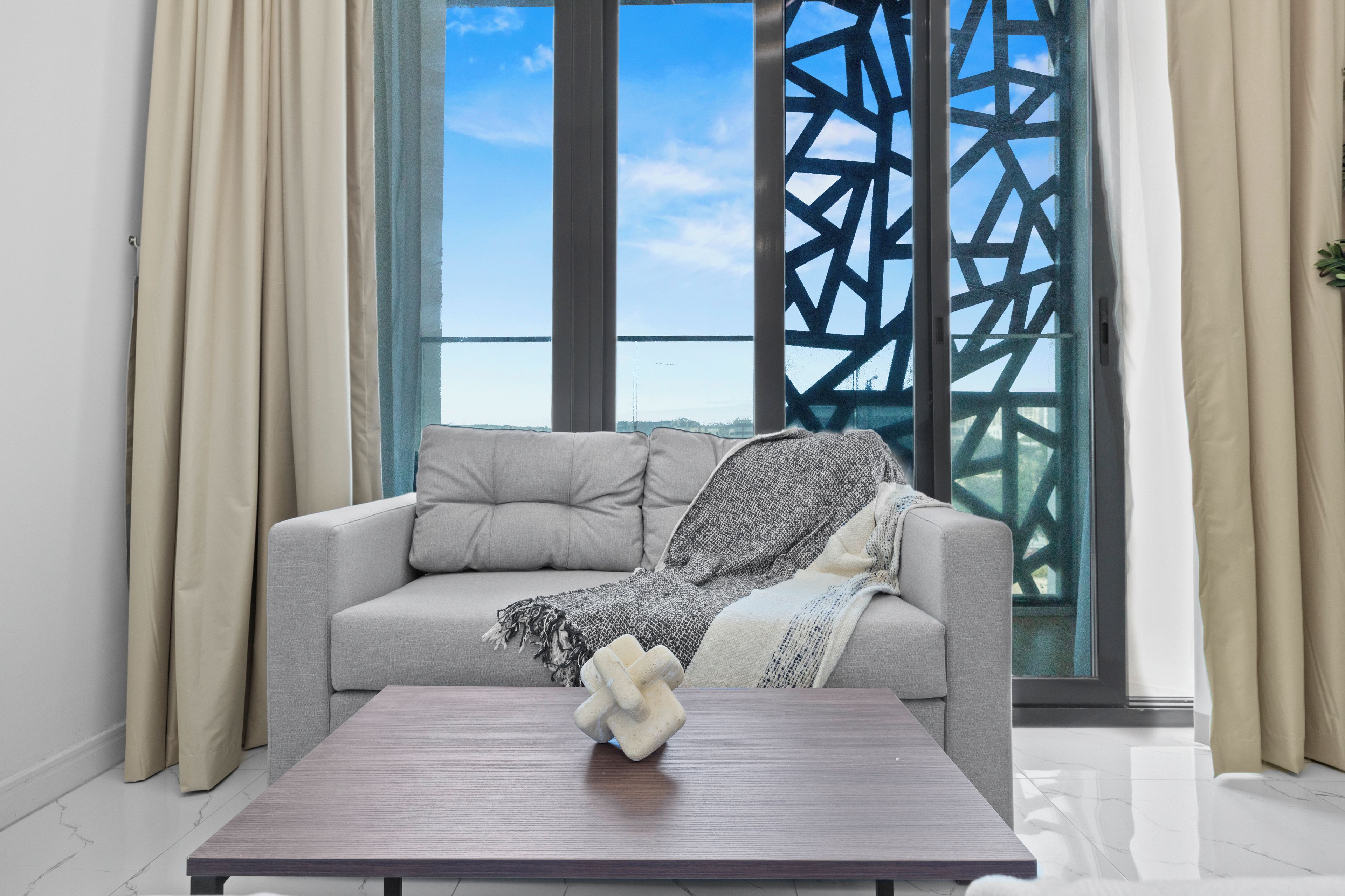 Ein modernes Wohnzimmer mit einem grauen Sofa, einem Holztisch und Meerblick durch ein Fenster mit geometrischem Muster.