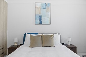 Une chambre à coucher comprenant un lit, deux tables de chevet, un tableau abstrait accroché au mur et une petite table de nuit avec une lampe.