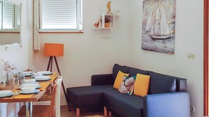 Un salon chaleureux avec un canapé bleu foncé, une table à manger en bois dressée pour deux personnes, une petite table d’appoint avec une lampe et un tableau représentant un voilier accroché au mur.