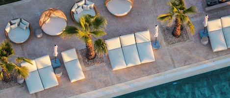 An aerial view of a poolside area with sun loungers, palm trees, and a flower bed.