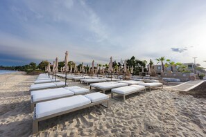 Un espace de détente aménagé sur la plage, avec des chaises longues et des parasols, un ciel dégagé et un bâtiment en arrière-plan.