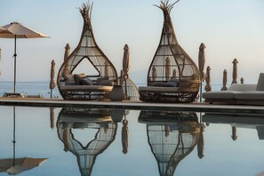 Deux fauteuils de relaxation en osier avec des coussins, placés au bord d’une piscine, un canapé blanc et un parasol de plage.