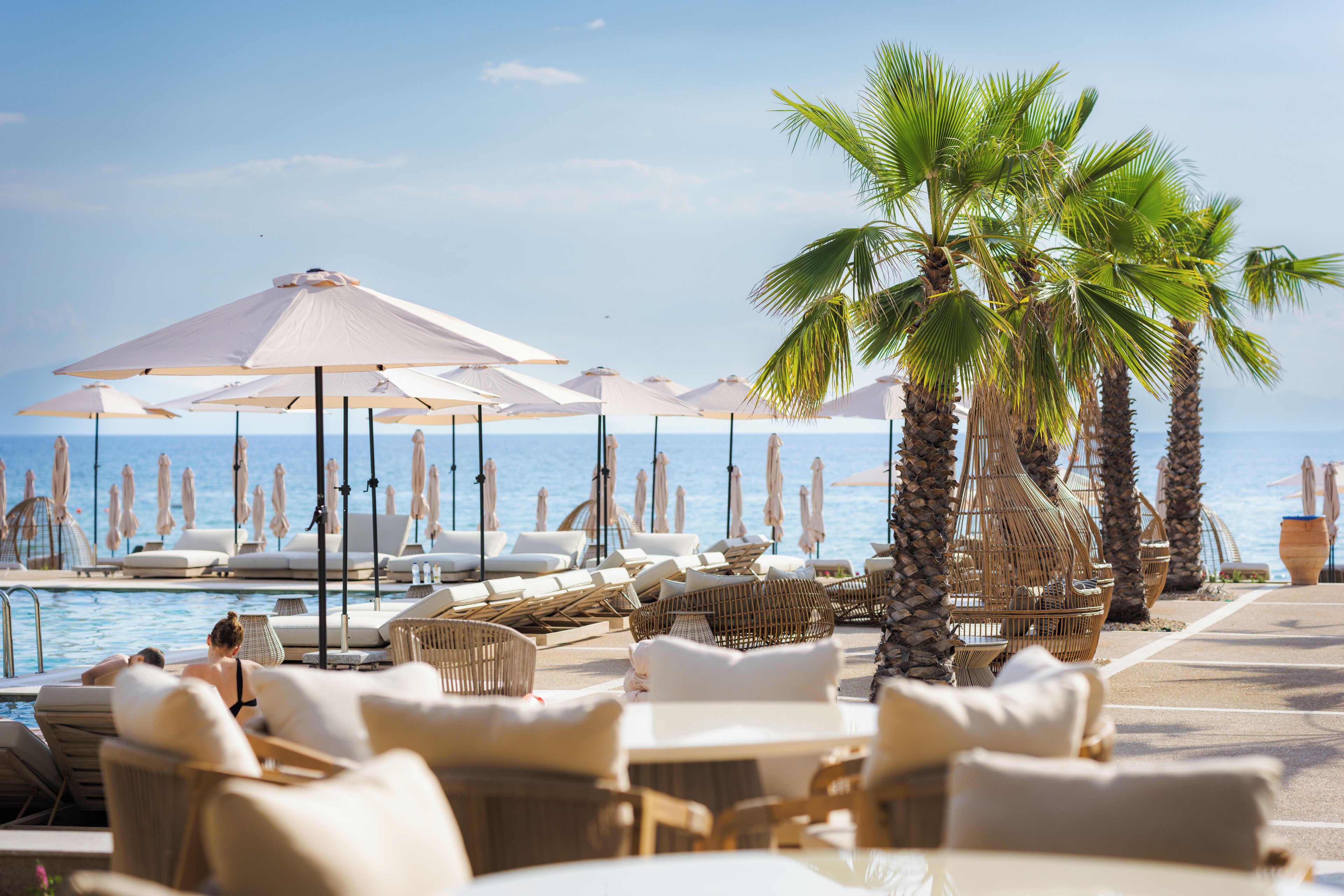 Un espace aménagé au bord de la piscine, avec des chaises longues et des parasols, des palmiers et une vue sur la mer.