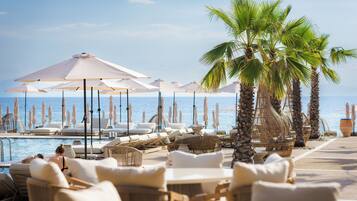 Un espace aménagé au bord de la piscine, avec des chaises longues et des parasols, des palmiers et une vue sur la mer.