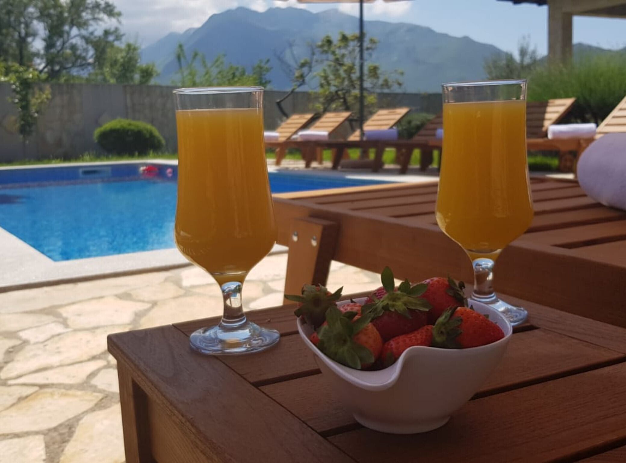 Due bicchieri di succo d'arancia su un tavolo di legno, con una ciotola di fragole, vicino a una piscina con lettini e montagne sullo sfondo.